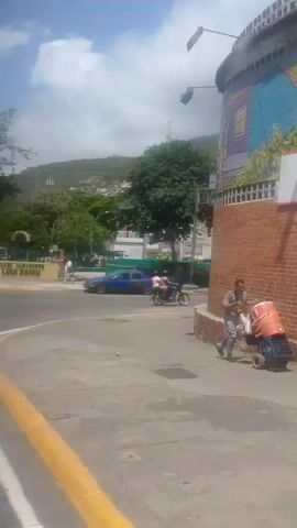 The video captures an outdoor scene with a street and a hillside in the background. A blue sedan is parked on the side of the road. A person in a yellow shirt is standing near the blue car. In the foreground, a person with gray hair wearing a green shirt is visible from the back. There is also a glimpse of another person's shoulder in the lower left corner. The environment appears to be a sunny day with trees and buildings on the hillside.