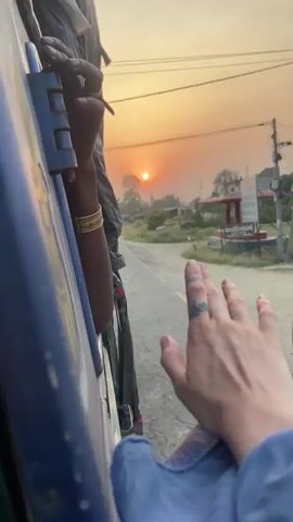 A person hangs out of a moving blue vehicle, their arm extended, hand open towards the passing scenery. Another person’s hand, adorned with gold bracelets, grips a handle on the vehicle frame. Two men on bicycles ride by on the asphalt road. Buildings, power lines, and vegetation line the roadside under a yellow-orange sky, presumably at sunrise or sunset. A blue auto-rickshaw passes by.