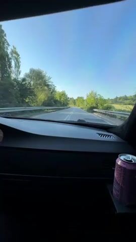 The video captures a first-person view from inside a car driving on a paved road during the daytime. The car's dashboard is visible in the foreground, with the windshield offering a clear view of the road ahead. A pink, textured can of what appears to be an energy drink is positioned in a cup holder on the right side of the dashboard. Text visible on the can includes "CELSIUS", "LIVE FIT", "SPARKLING ENERGY DRINK", and partially "MELON".

The car is moving along a two-lane road that is bordered by dense green trees and vegetation on both sides. The sky is bright blue and clear, indicating sunny weather. Around the 0:00:03 mark, a white circular road sign with a red border and the number "50" (likely denoting a 50 km/h speed limit) is briefly visible on the right side of the road. A screen on the car's dashboard shows the word "Navigation". The landscape suggests a rural or semi-rural setting, consistent with the Krivodol area in Bulgaria. The car continues to drive straight as the road gently curves.