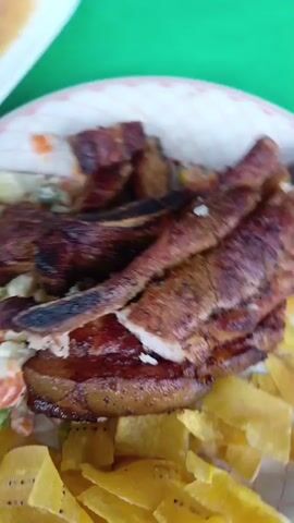 A plate of food is shown in a close-up, the plate is filled with fried plantain chips, salad, and grilled pork. The pork appears sliced and arranged on top of the other food items. A small dish containing what looks like beans is partially visible in the background. The overall setting appears to be casual, possibly a meal at a restaurant or outdoor event.