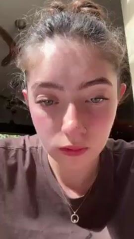 A close-up shot features a fair-skinned woman with light brown hair pulled up in a messy bun. Her face is front and center, revealing a nose ring, full pink lips, and green eyes. She is wearing a brown t-shirt and a delicate necklace with a circular pendant. The woman is applying makeup to her eyebrows with a brush, her expression serious and focused. Her fingernails are long and adorned with glittery polish. The background appears to be indoors, with a window casting natural light on her face. The overall focus is on the woman's face as she meticulously applies her eyebrow makeup.