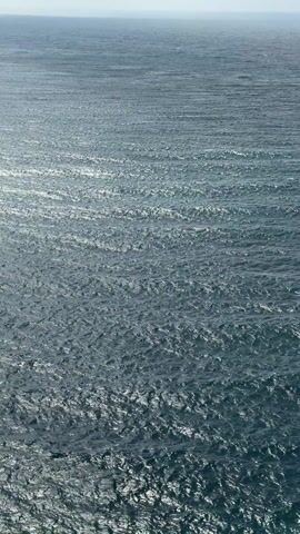 The video captures a wide, expansive view of a large body of water, most likely an ocean or a vast sea, under bright daylight. The water itself is a deep, somewhat muted blue-gray color. Its surface is not calm; instead, it is covered in numerous small, choppy waves that create a textured, shimmering appearance. Bright sunlight reflects off these wave crests, creating countless glinting and sparkling points across the water's surface, particularly in the middle and foreground of the frame. These reflections give the water an almost metallic or iridescent quality in places.

In the distance, a faint, light blue horizon line separates the water from a hazy, light-colored sky that occupies the top portion of the vertical frame. There are no landmasses, boats, people, or any other distinct objects visible in the scene, emphasizing the vastness and open nature of the water. The overall impression is one of natural beauty and tranquility, with the constant, subtle movement of the water being the main focus. The video is static, maintaining a consistent high-angle perspective looking out over the water.