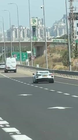 The video shows a white Chevrolet Corvette driving on a highway in Israel. The Corvette has a yellow license plate. In the background, there are signs in Hebrew and English indicating directions to Yakum. Other vehicles, including a white van, are also visible on the highway. The road has white lane markings and a metal barrier on the side. Buildings and some vegetation are visible in the distance. The sky is bright, suggesting it is daytime.