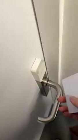 A person in a green sweater and blue jeans opens a white door with a silver handle using a key card. The door opens into a room with gray carpet and a blue wall with white trim. A red hat is visible on a surface behind the door. The person appears to be entering the room.