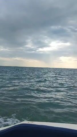 The video shows a view from the front of a boat moving through the ocean. The water is choppy with small waves. The sky is cloudy with patches of sunlight breaking through. The horizon line is visible, separating the water from the sky. The lower part of the boat's railing is visible in the foreground.
