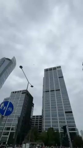 The camera looks up at modern buildings in Warsaw, Poland, on a cloudy day. A circular blue street sign showing a person walking and a person riding a bicycle is in the left foreground. A street lamp is also visible. In the distance, a small airplane flies across the sky.