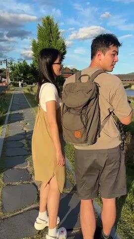 This video opens with a close-up shot of a woman wearing glasses and a tan overall dress over a white t-shirt, standing next to a man from the shoulders down, who is wearing a backpack. The camera then pans to the right, showing a paved walkway made of flat stones. To the left of the walkway is a road with a yellow auto-rickshaw driving by. There are planters with red flowers and greenery along the road and walkway. Further down the walkway, there are more trees and a bridge with traditional architecture over a body of water. The sky is blue with scattered clouds, and the lighting suggests it is late afternoon or early evening. The video focuses on the environment and the people as they walk along the path.