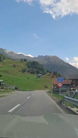 This video, filmed from a vehicle, captures a scenic drive through a mountainous landscape in Disentis/Mustér, Switzerland. The time of day appears to be late afternoon, as indicated by the bright sunlight and the shadows cast on the mountainside. The weather is clear with a bright blue sky and scattered white clouds.

The scene is dominated by a steep, grassy mountainside covered in lush green vegetation and dotted with scattered trees and a few small buildings. A winding asphalt road, marked with white lines, curves through the landscape, suggesting it's a mountain pass or a rural route. A metal guardrail runs along the side of the road, separating it from the steep incline.

In the distance, imposing, rugged mountains rise majestically, their peaks partially obscured by shadows. The overall impression is one of natural beauty and tranquility, with a sense of open space and fresh air.

There is visible text on a building to the right of the road, including what appears to be "REKORD," "SUPER MCD," and "SCHMIEDEMEISTER." A red and white circular sign with a number inside is also visible, likely a speed limit sign. Another sign with the word "BEER" and a web address is also present. The camera's perspective suggests the journey is ongoing, moving forward along the road.