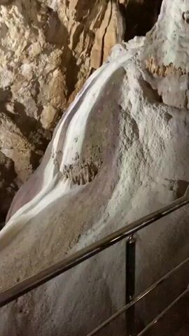 The video shows a cave. It begins with a close-up of a white, stone formation with a rough texture. A metal railing runs along the side. The camera then pans upward, showing a dark, rocky ceiling. In the distance, a light illuminates a section of the cave. The camera moves to a dark pathway where two people are walking. One person is wearing a tan outfit and a white hat, and the other person is wearing a white hoodie. The video ends with a shot of the cave floor, which is covered in dirt and rocks. The cave ceiling is visible above.