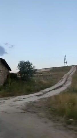 The video pans across a rural landscape. On the left, a stone building with an open window is visible, with a large stone slab leaning against it. The camera then moves to show the side of a stone building with a small window and an open doorway. A cross is visible on the roof. Further along, another stone building with a covered structure in front of it is seen, surrounded by overgrown vegetation. A power line tower stands on a hill in the background. The ground in the foreground appears to be a dirt or gravel road. The sky is overcast and pale blue.