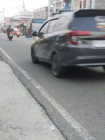 A dark gray SUV is driving on a paved road. There are multiple motorcycles on the road as well, some with riders and some without. The road appears to be in a town or city, with buildings and a sidewalk visible on the right side. There are also power lines and streetlights. The video appears to be shot from a low angle, close to the ground. The lighting suggests it is daytime, possibly late afternoon or early evening.