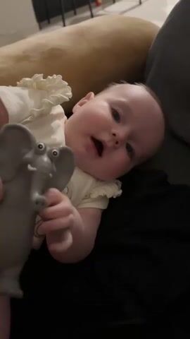 A baby is lying on their back, looking towards the camera. The baby is wearing a white onesie with ruffles on the sleeves and a picture of a teddy bear on the front. The baby is holding a gray elephant toy with googly eyes. The baby's left arm is raised and the toy is near their face. The baby's right arm is bent at the elbow and the hand is open. The baby's legs are bent at the knees and the feet are visible. The baby appears to be on a couch or other upholstered furniture. The background is blurred. The video is about 4 seconds long and shows the baby moving the toy.