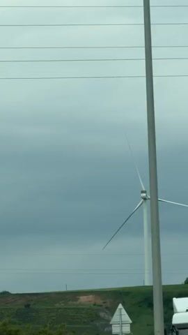 The camera follows a highway view from a moving vehicle. In the foreground is the top of a white truck that has “KÖKSAL ULUSLARARASI NAKLIYAT” printed on the side. In the background is a landscape view featuring a single wind turbine set against a gray sky. Power lines are visible at the top of the frame and along the horizon line in the distance. As the vehicle moves, objects such as a pole, a traffic sign, and trees pass in front of the turbine, briefly obscuring the view. Turkish music plays in the background.