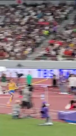 A male pole vaulter wearing blue and yellow athletic gear is performing a jump. He sprints toward the pole vault standard, plants the pole, vaults into the air, and attempts to clear the bar. He almost clears the bar, but slightly grazes it before landing on the mat. A crowd fills the stadium. There are several people on the field. The ground is red. The poles are lavender. The standard has various advertisements. A camera on a tripod is present.