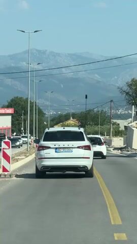 A white Skoda SUV is driving down a paved road, followed by another white SUV. The road has a yellow line marking the lane. On the left side of the road, there are streetlights, trees, and a building with red and white signage. In the background, there are mountains under a clear blue sky. The video is captured from inside another vehicle.