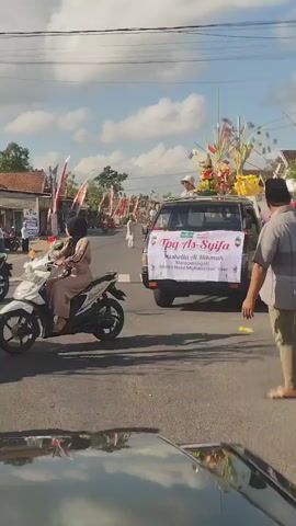 A black pickup truck drives down a paved road under a cloudy sky. The truck bed is adorned with decorative elements and carries several children wearing hijabs and some adult women. A banner on the front of the truck reads "TPA As-Syiifa Memperingati Maulid Nabi Muhammad Saw". A person on a motorcycle, also wearing a hijab and an orange dress, drives past the truck. Several Indonesian flags are visible along the side of the road. The overall scene appears to be part of a parade or procession.