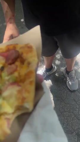 A person in a black t-shirt with a Mexican flag patch on the sleeve is holding a piece of pizza on a brown paper square. The pizza has a square crust and is topped with cheese, sliced sausage, and green bell peppers. The person is walking on a sidewalk, with shops and trees in the background. The person pauses and looks at the camera. The t-shirt has a barber pole design printed on it.