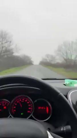 A driver in a car is going down a road in foggy, overcast conditions. The car's dashboard is visible, showing a speedometer reading around 80 km/h and a mileage of 10712 km. Trees line the road on either side. The driver's hands are on the steering wheel. The song "Children of the Revolution" by T. Rex is playing.