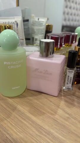 The video shows a close-up shot of several cosmetic products displayed on a wooden surface, likely a vanity or dressing table. In the foreground, there are three prominent bottles. On the left is a light green, frosted glass bottle with a rounded green cap, labeled "PISTACHIO CRUSH" and "KOTON". To its right is a rectangular, pale pink bottle with a silver cap, labeled "Miss Dior" and "LAIT CORPS RECONFORTANT NOURISHING BODY MILK ROSE". Further to the right is a small, clear glass rollerball bottle with a black cap, labeled "Gold" and "CREED AVENTUS". In the background, several other cosmetic items are visible, including tubes and bottles, some with gold accents. The overall impression is of a collection of beauty products arranged for display. The lighting is soft, and the focus is on the items in the foreground. The video appears to be a static shot with very minimal, if any, camera movement.