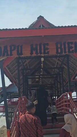 The video shows a group of people walking up a red wooden staircase toward a building with a thatched roof. The building has a sign above the entrance that reads "DAPU MIE BIE". The people appear to be entering a restaurant or food establishment. The environment is outdoors and appears to be a warm, sunny day.