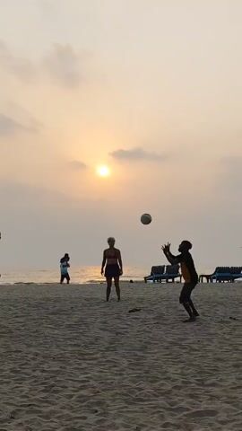 The video captures a group of people playing with a ball on a sandy beach. The sun is setting, and the sky is bright with a hazy appearance. There are four people prominently visible in the foreground. Two are men wearing swim trunks, and one is a woman wearing a sports bra and shorts. All appear to be playing with the ball. There are more people visible on the beach, including some walking along the shoreline and some relaxing on beach chairs. The sand is a light tan color, and the ocean is calm.