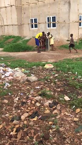The image captures an outdoor scene in Isieke, Nigeria, during the daytime. In the foreground, a large pile of assorted garbage, including plastic bags, bottles, and other waste, is scattered across a patch of dry grass and dirt.

In the midground, a group of approximately six individuals, consisting of one adult male and several children, are gathered around a water source, possibly a communal faucet or pump. The adult male, wearing a yellow shirt and blue shorts, stands near the water source, seemingly assisting the children. Some of the children appear to be partially undressed, possibly engaging in washing or bathing. One child in a brown shirt walks away to the right. The ground in this area is mostly dirt with some sparse green vegetation.

In the background, a light-colored, multi-story building with several white-framed windows is visible. Bamboo scaffolding is erected along the side of the building, suggesting ongoing construction or renovation. The immediate area around the building also features dirt and some greenery. The sky is not visible, but the lighting indicates it is daytime. No discernible text is visible in the image.