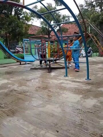 The video shows a playground with children on and around the play equipment. There's a red-roofed building partially visible in the background, along with trees. The ground is concrete and appears wet.  A metal structure resembling a climbing frame with a rotating element is the focal point. A child in a blue patterned shirt and pants stands next to it. Another child is already on the rotating part. Other children stand around. A slide and other play structures are visible in the background.  The children appear to be playing and talking. The video is somewhat shaky.