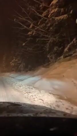 The video is taken from the dashboard of a car driving on a snow-covered road at night. The headlights illuminate the road ahead, revealing packed snow and tire tracks. Dense, snow-laden evergreen trees line both sides of the road, creating a dark and enclosed environment. There is a distant amber light visible on the left side of the road. The dashboard is visible in the foreground, showing some frost or condensation. The overall scene depicts a wintery and possibly remote setting, with the car navigating the snowy road under low-light conditions.