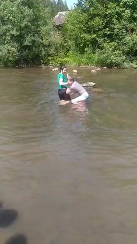 The video shows two young people in a shallow river on a sunny day. The person on the left is wearing a green t-shirt and dark shorts, and is standing and facing the other person. The person on the right is wearing a grey t-shirt and light-colored shorts, and is partially submerged in the water, facing the person on the left. They appear to be interacting, possibly playing or splashing each other, as there is disturbance in the water around the person on the right. The riverbed is visible in places, with some rocks. The background consists of lush green trees and vegetation on the riverbanks, and a building structure with a roof is visible further back through the trees. The water is murky brown.