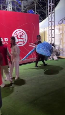 A person in a red and brown costume is carrying a large blue stuffed creature with a swirled pattern. The person is walking in front of a white tent with arched windows. To the left of the frame is a large red panel with a logo. There are people in the background and to the right of the frame, some of whom are looking at the person. The ground is covered with green artificial turf. The video appears to be shot at night or in a dimly lit area.