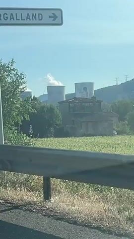 The video shows a moving shot from a vehicle, likely on a road with a guardrail in the foreground. The scenery includes trees and vegetation. At one point, large industrial cooling towers with plumes of steam are visible in the distance. Later, a road sign displaying "50 RAPEL VILLAGE PRUDENT" appears. The sky is clear and blue.