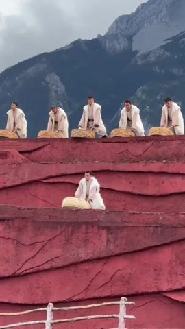 Six men in white robes are playing drums. Five of them are standing on a raised platform, while one man is positioned below them on a lower platform. The drums are large and cylindrical, and the men are using drumsticks to play them. The background shows a mountain range with a cloudy sky. The platforms appear to be made of a rough, reddish-brown material, possibly rock or sculpted earth. The man in the foreground is looking down at his drum as he plays, while the men on the upper platform are looking forward, presumably at an audience or a conductor. The overall scene suggests a performance or ceremony taking place outdoors.