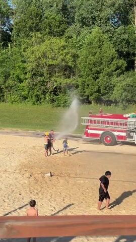 A fire truck with the number 3 painted on its side is parked on a sandy surface. A man is holding a hose connected to the fire truck and spraying water towards the left. Two young boys are running in the sand. One boy is in the foreground, facing away from the camera, and the other is in the background, also facing away. A third boy is standing near the man holding the hose. The background consists of a grassy slope and a dense green forest. The sun is shining, casting shadows on the sand. The scene appears to be a recreational area or park on a sunny day.