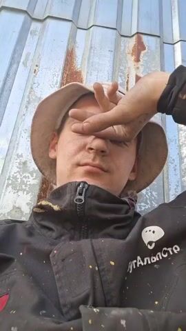 A man with a hat and dirty clothes is lying down with a hand shielding his eyes. He sticks his tongue out and then closes his mouth. He is lying near some corrugated metal. The man's black jacket has some spots of paint on it and the word "УГАЛЬПСТО" is printed in white.