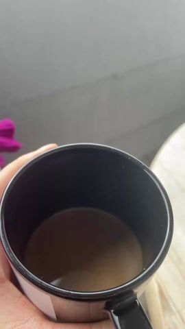 A hand holding a black mug containing a light brown liquid is positioned in front of a concrete balcony wall. Beyond the wall is a view of a suburban landscape consisting primarily of houses with red-tiled roofs. The sky is overcast with a mix of gray and white clouds, with patches of blue visible. The camera tilts upward to fully capture the cloudy sky.