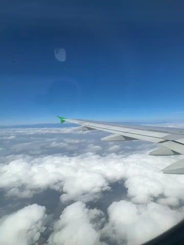 The video shows a view from an airplane window, looking out at the sky and clouds. The airplane's wing, with a green tip, is prominently visible in the foreground, extending across the frame. Below the wing, a vast expanse of white, fluffy clouds stretches towards the horizon. In the distance, hazy mountain ranges are visible peeking through the cloud cover. The sky is a deep, clear blue, suggesting good weather and a bright day. The overall scene evokes a sense of travel and the peacefulness of being above the clouds. There are no people visible. The video captures a moment in time during an airplane flight, with the focus on the aerial landscape. The time of day appears to be daytime, given the bright sunlight illuminating the clouds and sky. The location is Pesquería, Mexico, as indicated by the context.