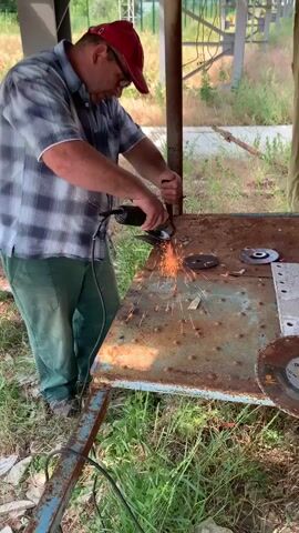 A man wearing a red baseball cap, glasses, a blue plaid shirt, and green pants is standing and using an angle grinder to cut a metal object on a rusty workbench. Sparks are flying from the angle grinder. The workbench appears to be outdoors, with grass and weeds growing around it. There are several metal discs on the workbench. The man is holding the metal object with his left hand and the angle grinder with his right hand. He is focused on the task at hand.
