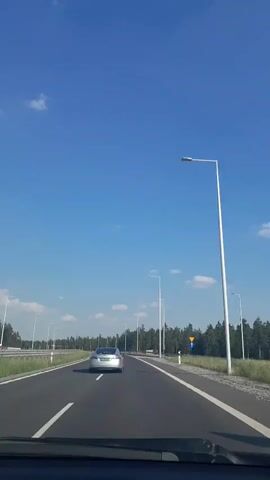 This is a vertical, first-person perspective video clip captured from inside a vehicle traveling on a highway. The scene is set on a multi-lane road bordered by a dense forest on the right and a lighter green grassy area with trees on the left. The sky is a clear, bright blue with a few scattered white clouds, suggesting a pleasant day. Tall, white streetlights line the highway at regular intervals.

A silver car is visible in front of the camera vehicle, traveling in the same direction. The road markings, including white solid and dashed lines, are clearly visible, indicating the lane structure. On the right side of the road, a yellow triangular warning sign with a black symbol is visible, mounted on a white post. The sun appears to be high in the sky, casting shadows, indicating it is likely midday. The overall impression is one of a smooth, uneventful journey on a well-maintained highway, likely in a rural or semi-rural setting, given the extensive tree line. The video quality is average, with some minor shakiness typical of footage recorded from a moving vehicle. No text is visible in the clip.