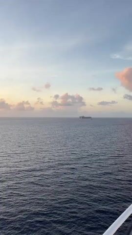 The video depicts a cruise ship sailing on a vast expanse of ocean under a partly cloudy sky. The sky exhibits hues of blue and light pink, suggesting either sunrise or sunset. The ocean is deep blue with gentle ripples on the surface. The cruise ship appears relatively small in the distance, emphasizing the vastness of the ocean. The camera is static, suggesting it is positioned on another vessel or elevated platform. The overall scene conveys tranquility.