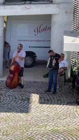 The video captures an outdoor performance by a three-piece band in Mojkovac, Montenegro. In the foreground, a man plays a double bass while singing into a microphone. To his right, another man plays an accordion. Seated behind them, a third man watches the performance.

The band is set against a backdrop featuring a white van with "Katuša" written in cursive script and an email address "sirankatunjanja@gmail.com". The ground is paved with cobblestones, and sunlight suggests it is daytime, likely warm weather. The overall atmosphere appears relaxed and informal, with the musicians seemingly enjoying their performance. The presence of the van and the surrounding architecture hint at a public space, possibly a street or a courtyard area. The subjects are dressed casually, indicating a leisurely event.