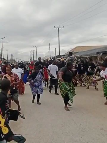 A group of people are walking down a street. Some are dancing and singing. The sky is cloudy and there are buildings on either side of the street.