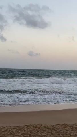 The video shows a view of a beach and the ocean. The sand in the foreground is light brown with visible texture. The ocean is a greenish-blue color with white waves breaking and rolling towards the shore. The sky above is a pale blue with scattered clouds, some of which are darker gray. The lighting suggests it is either early morning or late evening. There are no people or man-made objects visible in the frame.