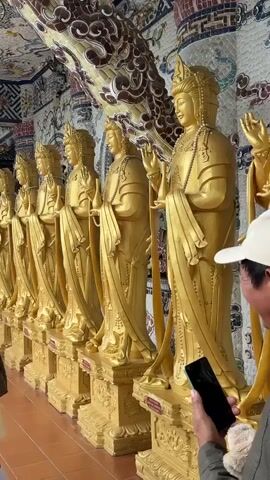 The video displays a row of golden statues, possibly deities or important figures, arranged in a line. A young girl with dark hair, wearing a light-colored dress and a brown jacket, stands in front of the statues, looking at the camera and smiling. To her left, a man in a black leather jacket is visible, looking down at his phone. Another person's hand holding a phone is visible in the lower right corner of the frame, seemingly capturing the scene. The background features an intricately decorated wall or ceiling with mosaic-like patterns in various colors, including white, blue, red, and gold. The lighting appears to be interior, possibly within a temple or a similar sacred space. The statues themselves are tall and adorned with ornate headwear and jewelry.