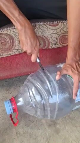 A close-up shot shows a person's hands holding a large plastic water jug that is partially filled with water. The person is using a knife to cut into the side of the jug. The jug is resting on a dirt or concrete surface, and part of a red upholstered couch is visible in the background. The person is wearing a white tank top and black pants.
