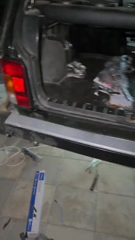 The video shows the back of a black car, possibly an SUV or station wagon, with the trunk open. The trunk appears to be empty and somewhat dirty, with some debris visible on the floor. A pink bucket is located on the right side of the trunk. Outside the trunk, on the ground, there are various tools and objects, including a metal rod, a roll of wire, a toolbox, and what appears to be a bottle of liquid. The ground is paved with square tiles. The lighting suggests it is either nighttime or indoors with artificial lighting. In some frames, a person's feet, wearing sandals, are visible at the bottom of the screen, suggesting the person filming is standing near the car. The scene appears to be a workspace or garage area.