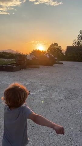 A young boy with blond hair is running towards the sunset in a gravelly area. He is wearing a gray t-shirt and green shorts with black and white striped socks and sandals. In front of him are two large metal dumpsters filled with what appears to be trash. A smaller metal container is to the left. The sun is setting in the distance, casting a warm orange glow across the sky and silhouetting the mountains and trees.