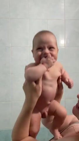 A close-up shot shows a baby being held up by two hands against a light green tiled wall. The baby is naked and appears to be laughing or making a funny face, with its right hand in its mouth. The baby is wearing a thin red string bracelet on its left wrist. The hands holding the baby are out of focus. The lighting is bright, and there is a white light reflection in the upper right corner of the wall.