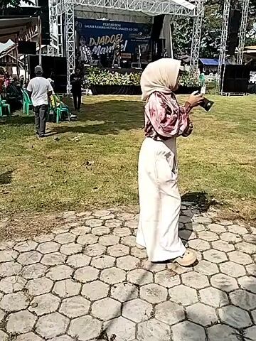 A video shows an outdoor scene with a stage, a grassy field, and a group of children in marching band uniforms. A woman wearing a hijab and carrying a camera walks towards the stage. A man in sunglasses walks past her. The camera pans to show a line of children holding drums, dressed in black and red marching band uniforms with hats. Another child stands out in a colorful green and pink costume. The children are standing on a grassy field with a tiled walkway. In the background, there are buildings and people walking around.