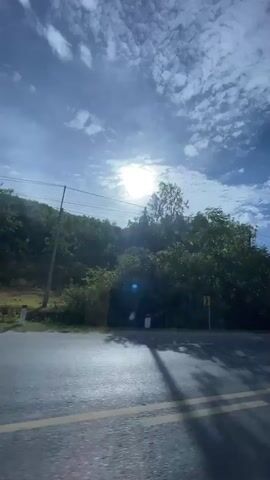 The video shows a road with a yellow line down the center, bordered by green trees and hills on one side. The sun is shining brightly through the clouds, casting shadows on the road. In the first few seconds, a motorcycle can be seen riding down the road, carrying bags on its back. The camera appears to be moving, giving a sense of travel along the road. The environment seems to be rural, with vegetation and a clear sky.