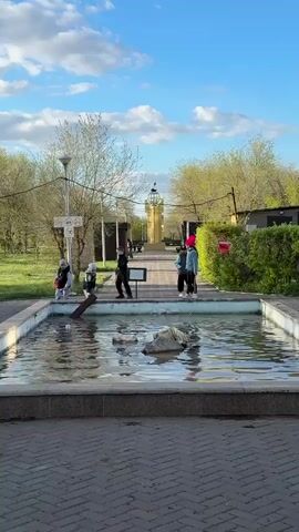 A group of people, including children, are gathered around a rectangular concrete fountain in a park on a sunny day. The fountain contains water and has some rocks in the center. A small sign stands next to the fountain. The people are mostly walking and standing near the fountain.  In the background, a brick walkway leads to a tall, golden, tower-like structure. The park has trees with light green foliage, and a few lampposts. The sky is blue with scattered clouds.